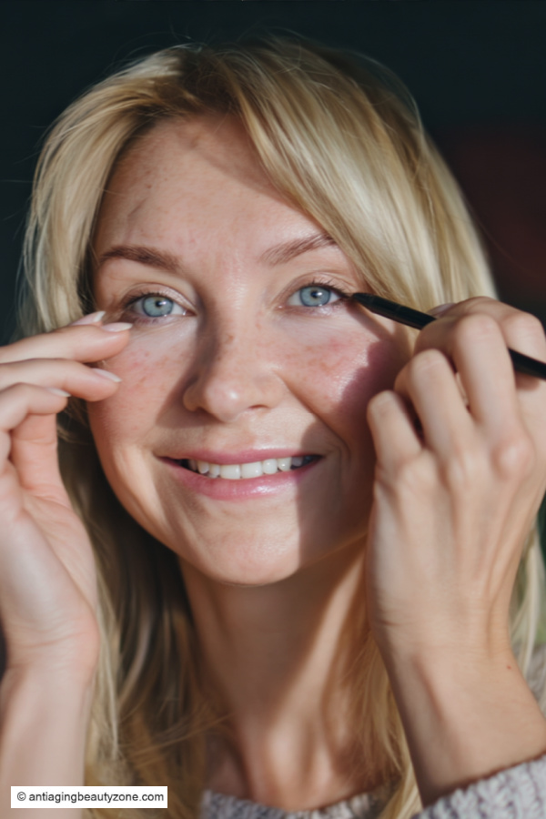 Woman applying pencil eyeliner to upper lash line on mature eyelids, showing soft definition for older eyes