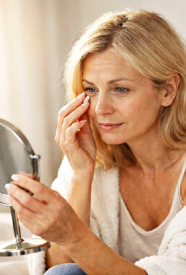 Woman noticing under-eye puffiness while looking in the mirror in soft morning light