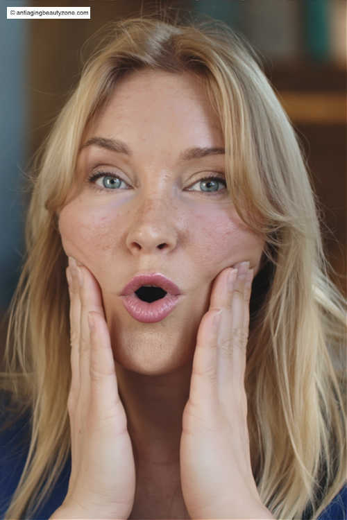 Mouth-corner lift: gentle lower-face move Woman making a soft “O” with hands at cheeks, demonstrating the mouth-corner lift for jowls.