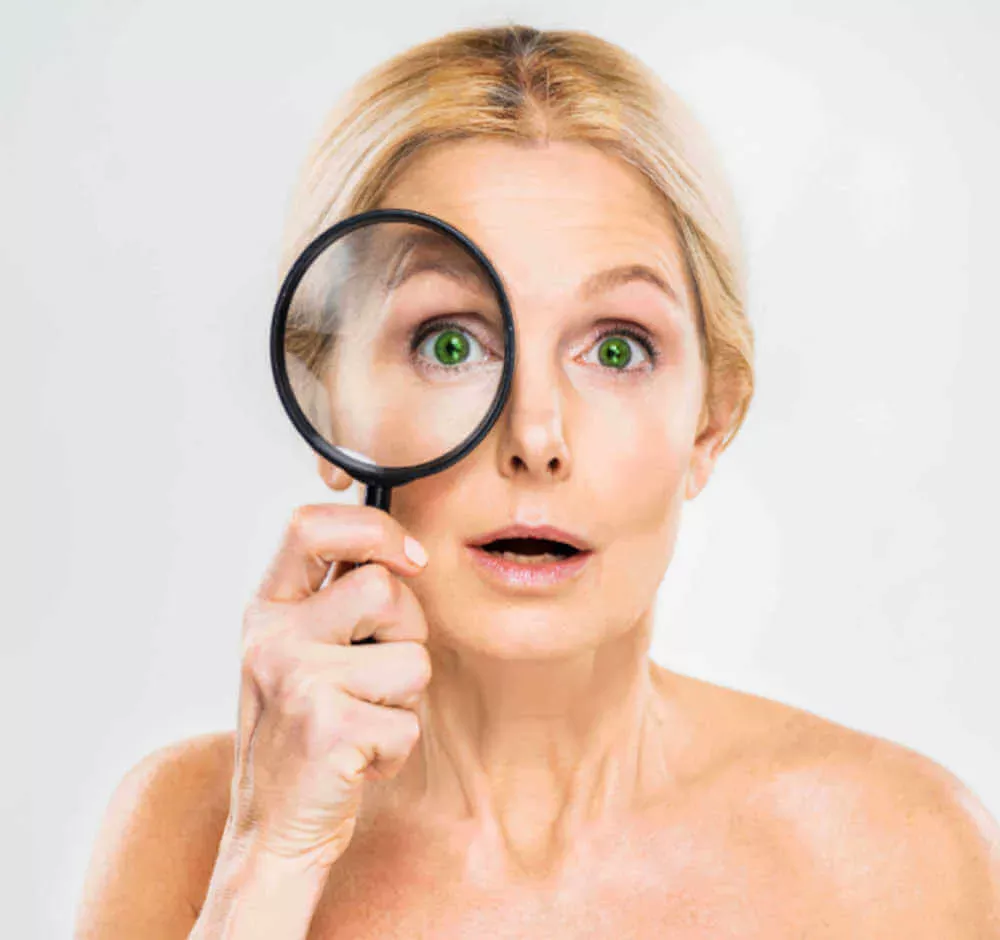 Surprised middle-aged woman examining her skin with a magnifying glass to check for wrinkles and fine lines.